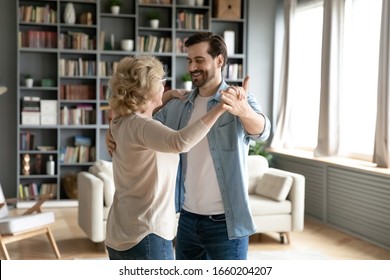 Happy Senior Woman Dancing With Grown-up Son Enjoy Family Weekend At Home Together, Smiling Middle-aged 70s Mother Waltz Sway With Adult Man Child, Relax In Living Room, Family Bonding Concept