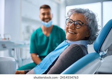 Happy senior woman at dental clinic looking at camera. Her dentist is in the background. - Powered by Shutterstock