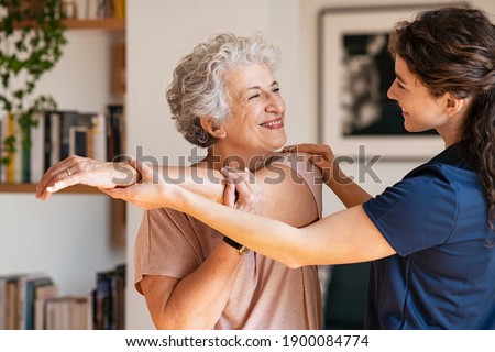 Happy senior woman doing exercise at home with physiotherapist. Old retired lady doing stretching arms at home with the help of a personal trainer during a rehabilitation session.