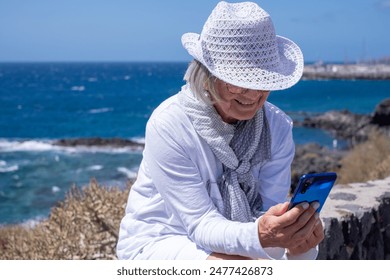 Happy Senior Woman dressed in white Sitting Outdoor in a Windy day Feeling in Vacation at Sea. Elderly Female Talking Smiling using Mobile Phone - Powered by Shutterstock