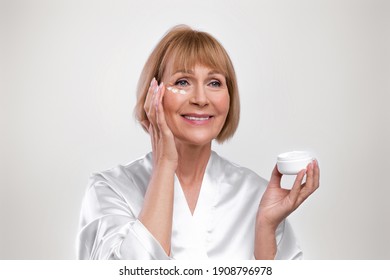 Happy senior woman in dressing gown applying anti-aging face cream on light studio background. Beautiful mature lady using rejuvenating skincare product, pampering herself