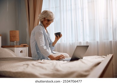 Happy senior woman drinking coffee and using laptop while relaxing in her bedroom in the morning. Copy space. - Powered by Shutterstock