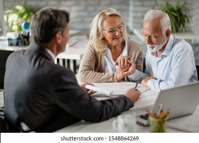 Happy senior woman encourages her husband to sign a contract with their bank manager during a meeting in the office. 