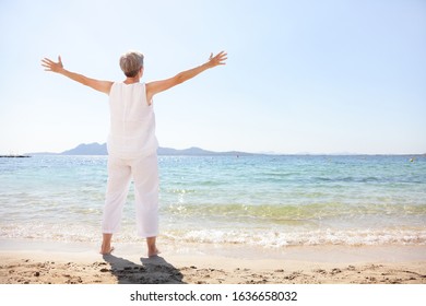Happy senior woman enjoying freedom of early retirement carefree on summer holiday in happiness standing with arms outstretched at beach. Full length view of lady tourist wearing linen clothing.