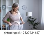 Happy senior woman examines her weight loss progress by wearing oversized pants. She stands in modern room, proud of healthy lifestyle and personal achievement in fitness journey.