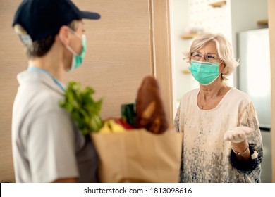 Happy Senior Woman And Female Courier Wearing Protective Face Masks While Communicating At The Doorway During Coronavirus Epidemic. 