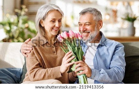 Happy senior woman getting romantic present from husband on Valentines Day while sitting on sofa at home, selective focus. Pleased elderly female receiving bouquet of spring flowers on March 8