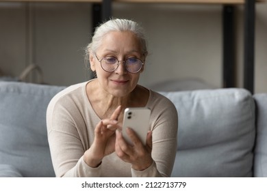 Happy senior woman in glasses reading text message on mobile phone, typing, browsing Internet, social media, using online ecommerce shopping app, making video call at home, sitting on couch