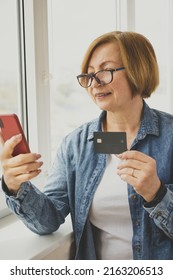 Happy Senior Woman Holding Bank Credit Card Using Smartphone, Making Internet Payment, Online Shopping, Booking Hotel, Order Taxi Via App, Standing Near Window Indoor. Online Banking.Vertical. Mockup