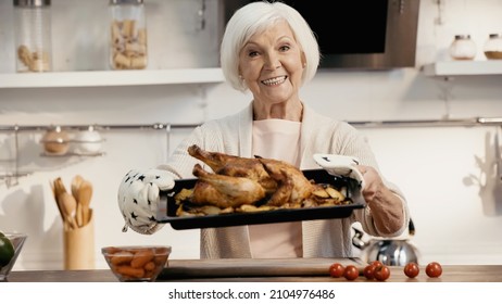 Happy Senior Woman Holding Oven Sheet With Roasted Turkey Near Fresh Cherry Tomatoes And Carrots In Kitchen