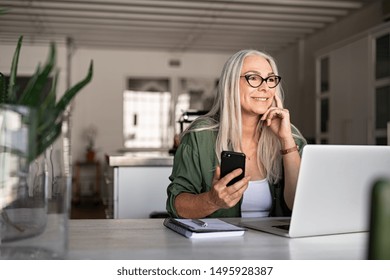 Happy Senior Woman Holding Smartphone And Laptop Daydreaming While Looking Away. Successful Stylish Old Woman Working At Home While Thinking. Fashionable Lady Entrepreneur Wearing Cool Eyeglasses.