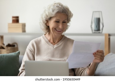 Happy Senior Woman With Laptop Reading Paper Letter At Home, Getting Good News And Smiling. Middle Aged 60s Lady Receiving Insurance Agreement Or Tax Document, Paying Bill, Reviewing Invitation