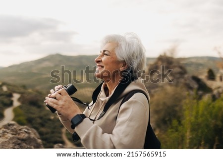 Happy senior woman looking at the view while standing on a hilltop with binoculars. Cheerful elderly woman enjoying a leisurely hike outdoors. Woman enjoying recreational activities after retirement.