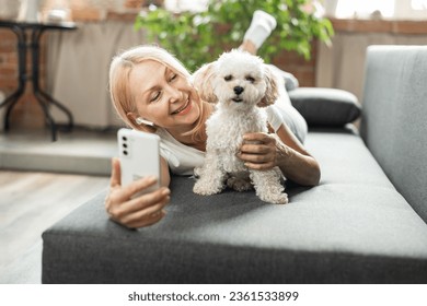 Happy senior woman lying on sofa, using phone and internet app for videocall while pet dog nearby. Concept of using gadgets and technology by older people - Powered by Shutterstock