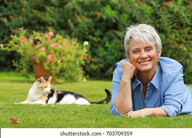 Happy Senior Woman Is Lying On The Grass In The Garden With A Cat In Background