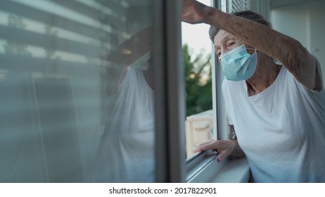 Happy Senior Woman In Mask Waves To Family From Window Hospital. Masked Elderly Female Glad That Children Came Visit Her In Nursing Home. Grandmother Greets Child With Hand Of Relatives At Clinic