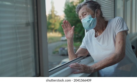 Happy Senior Woman In Mask Waves To Family From Window Hospital. Masked Elderly Female Glad That Children Came Visit Her In Nursing Home. Grandmother Greets Child With Hand Of Relatives At Clinic.