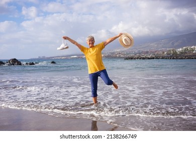 Happy senior woman with outstretched arms on sea shore holding shoes and hat in hands looking at camera. Elderly grandmother barefoot in the water enjoying vacation and freedom - Powered by Shutterstock