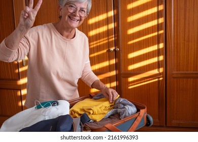 Happy Senior Woman Packing Her Stuff Into A Suitcase On The Bed Preparing For Summer Vacation Trip. Traveling Preparation Concept