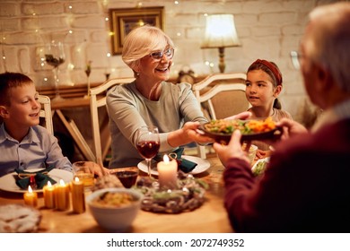 Happy Senior Woman Passing Food While Eating Thanksgiving Dinner With Her Family In Dining Room.