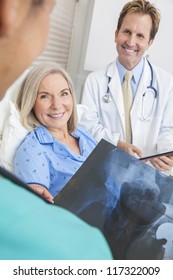 Happy Senior Woman Patient Recovering In Hospital Bed With Male Doctor And Female Nurse Looking At Hip Replacement X-ray