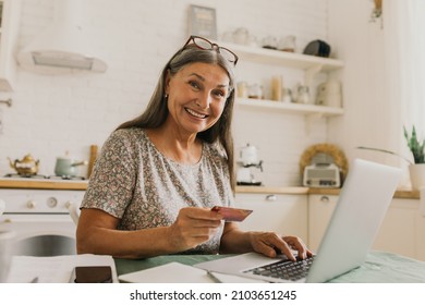 Happy Senior Woman Paying For Orders. Open Laptop With Information. Smiling Lady Sitting On Kitchen, Cozy Home Interior. Modern Technology, Online Shopping, Learn New Skills Concept