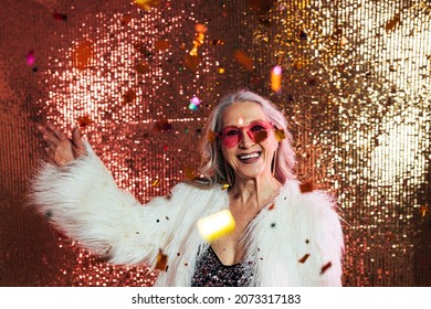 Happy Senior Woman In Pink Eyeglasses And White Fur Coat Having Fun Under Confetti