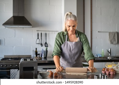 Happy Senior Woman Preparing Christmas Cookies At Home. Mature Woman Rolling Dough For Biscuits On Kitchen Counter. Old Lady Wearing Apron Rolling Pastry With Wooden Rolling Pin And Smiling.