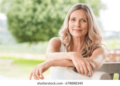 Happy Senior Woman Relaxing On Bench In The Lawn. Close Up Face Of A Mature Blonde Woman Smiling And Looking At Camera. Retired Woman In Casuals Sitting Outdoor In A Summer Day.