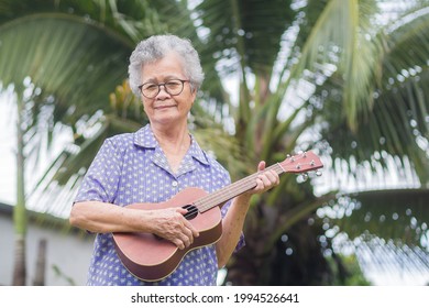 Happy senior woman with short gray hair playing the ukulele while standing in a garden with a coconut tree background. Enjoy life after retiring. Concept of aged people and relaxation.