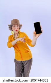 Happy Senior Woman Showing Empty Phone Screen Pointing To Phone On Light Grey Background With Blank Space For Text.