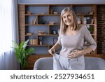 Happy senior woman showing successful weight loss journey while standing indoors holding oversized pants, symbolizing health, achievement, and positive lifestyle change.