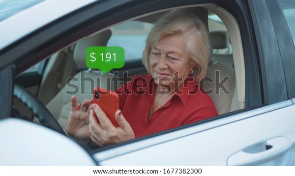 Happy Senior Woman Sits In Car Use Red Cell Phone\
Smile Businesswoman Blonde. App Icon with Online Transaction.\
Financial Transactions in the Smartphone. Receive a Message About\
Increase Money.