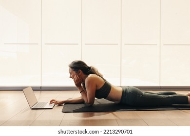 Happy Senior Woman Smiling While Joining An Online Yoga Class On A Laptop. Woman Following A Virtual Fitness Tutorial At Home. Cheerful Mature Woman Lying On An Exercise Mat.