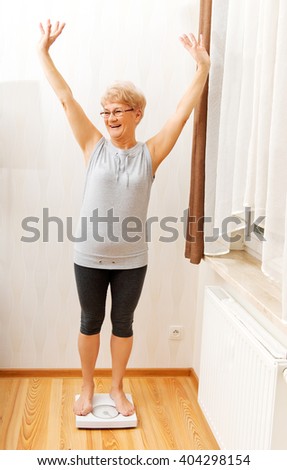 Happy senior woman standing on weight scale in living room
