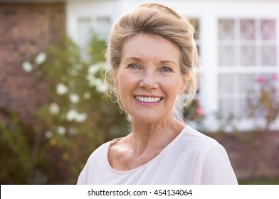 Happy senior woman standing outside her house. Content woman smiling and looking at camera. Portrait of cheerful old grandmother relaxing outside the house. - Powered by Shutterstock