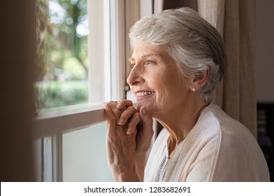 Happy Senior Woman Standing At Window And Looking Outside. Beautiful Grandmother Smiling While Looking Through The Window. Cheerful Old Woman Relaxing In Home While Standing At Window Sill.