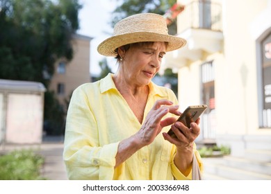 Happy Senior Woman In Sun Hat  Using Smartphone, Looking At Screen On Summer City