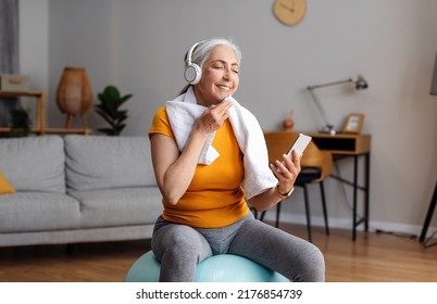 Happy Senior Woman With Towel On Shoulders And Wireless Headset Sitting On Fitball, Listening To Music On Cellphone, Resting During Home Workout, Free Space