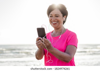 Happy Senior Woman Tracking Work Out Fitness From The Phone Outdoor. 
Sixsties Woman Wearing Headphones Doing Jogging At The Beach. Concept About Mature People, Sport And Technology. 