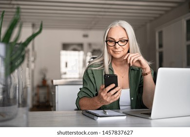 Happy senior woman using mobile phone while working at home with laptop. Smiling cool old woman wearing eyeglasses messaging with smartphone. Beautiful stylish elderly lady browsing site on cellphone. - Shutterstock ID 1457837627