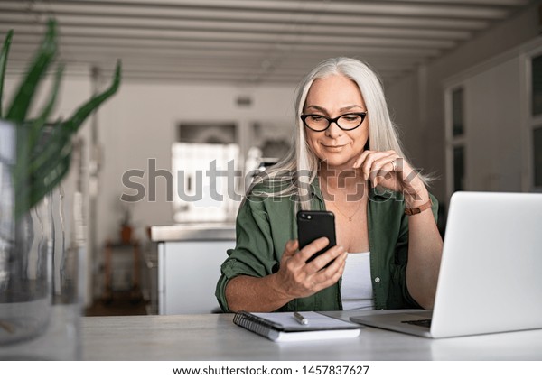 Happy senior woman using mobile phone while\
working at home with laptop. Smiling cool old woman wearing\
eyeglasses messaging with smartphone. Beautiful stylish elderly\
lady browsing site on\
cellphone.