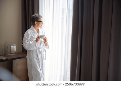 Happy senior woman in white bathrobe holding cup of tea, looking out the window in hotel - Powered by Shutterstock