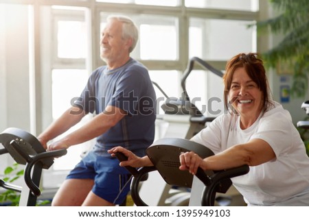 Happy senior woman working out in gym. Smiling elderly couple exercising in gym on stationary bicycle, focus on woman.