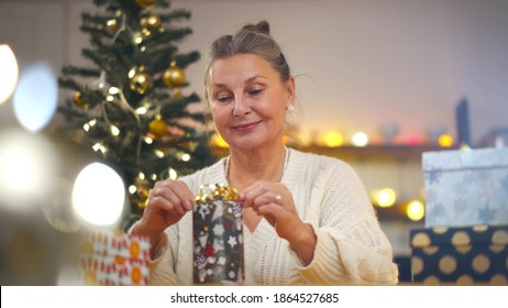 Happy senior woman wrapping christmas presents for grandchildren. Portrait of aged grandmother packing new year gift with decorated christmas tree on background - Powered by Shutterstock