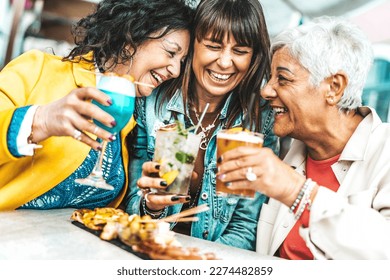 Happy senior women drinking cocktail glasses sitting at bar table - Group of best friends enjoying happy hour cheering drinks at pub restaurant - Life style concept with girls hanging out together - Powered by Shutterstock