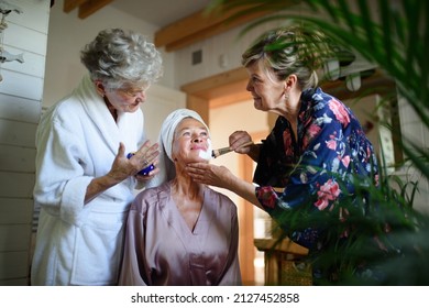 Happy Senior Women Friends In Bathrobes Applying Face Mask Indoors In Bathroom, Selfcare Concept.