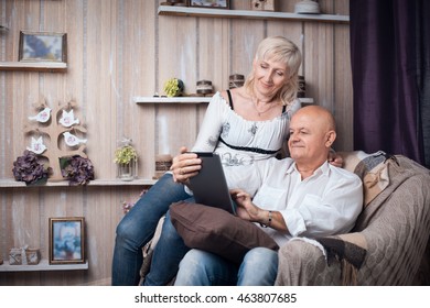 Happy Seniors Couple Reading From Tablet In Cozy Room; Elder Male And Female Browsing Internet With Mobile Device;