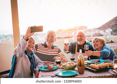 Happy seniors friends taking selfie with mobile smartphone camera at barbecue dinner - Mature people having fun eating and drinking red wine on patio while using new trends technology phone apps  - Powered by Shutterstock