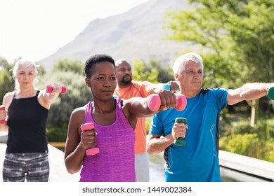 Happy Seniors And Mature Couples Exercising With Dumbbells. Healthy Multiethnic People Exercising Using Dumbbells Outdoor. African Couple And Senior Friends In Sportswear Stretching Arms At Park.